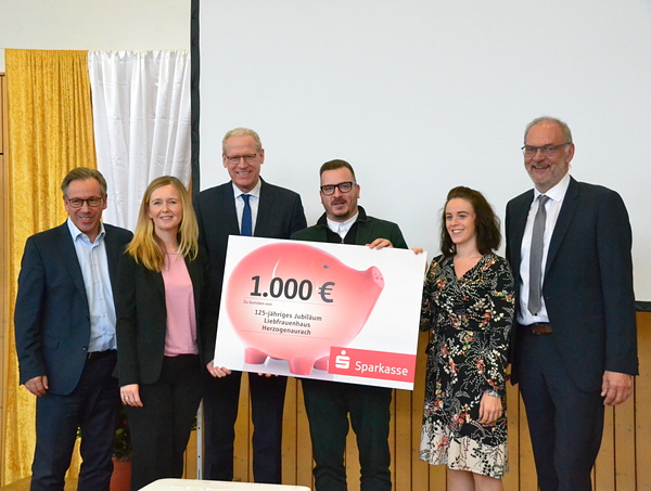 Auf der Bühne steht eine Gruppe Menschen, die einen symbolischen großen Spendenscheck halten mit einer Summe von 1.000 Euro